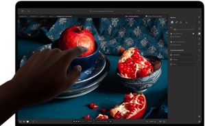 MacBook Pro mit PROLED XDR Display mit einem Stillleben mit Granatäpfeln, blauer Keramik und Textilien, eine Hand reicht nach dem Bildschirm, um mit dem Bild zu interagieren